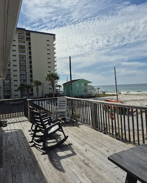 A wooden deck with chairs and a table, overlooking a beach with a high-rise building and a green house in the background.