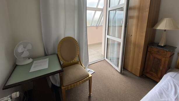 A hotel room with a desk, chair, and a bed. There is a window with curtains, a fan, and a lamp on a nightstand. - Studio Golden 2 (Hlavní mesto Praha)