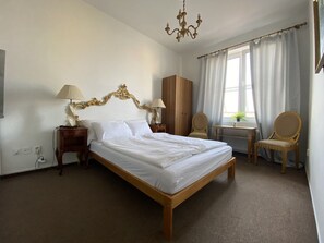 A hotel room with a large bed, two bedside tables, a chandelier, a wardrobe, a desk with a chair, and a window with curtains. - Studio golden (Hlavní město Praha)