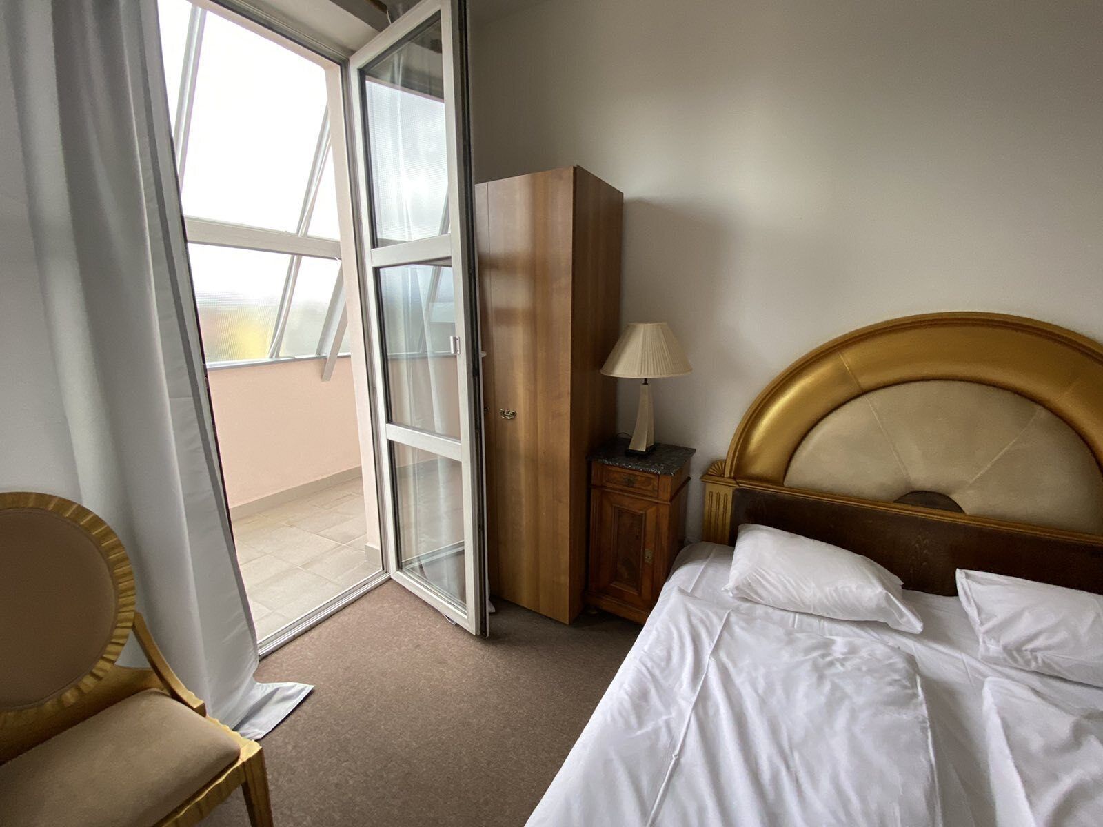 A hotel room with a bed, a nightstand, a lamp, a chair, and a sliding glass door leading to a balcony.