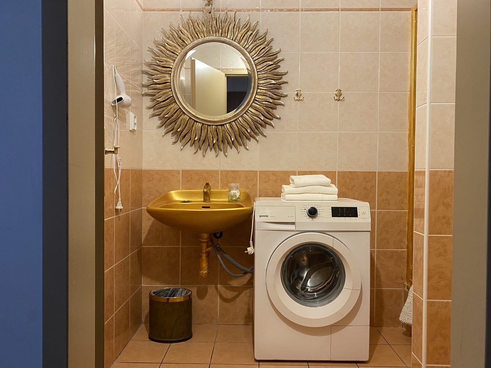 A bathroom with a golden sink, a washing machine, and a mirror with a sunburst frame.