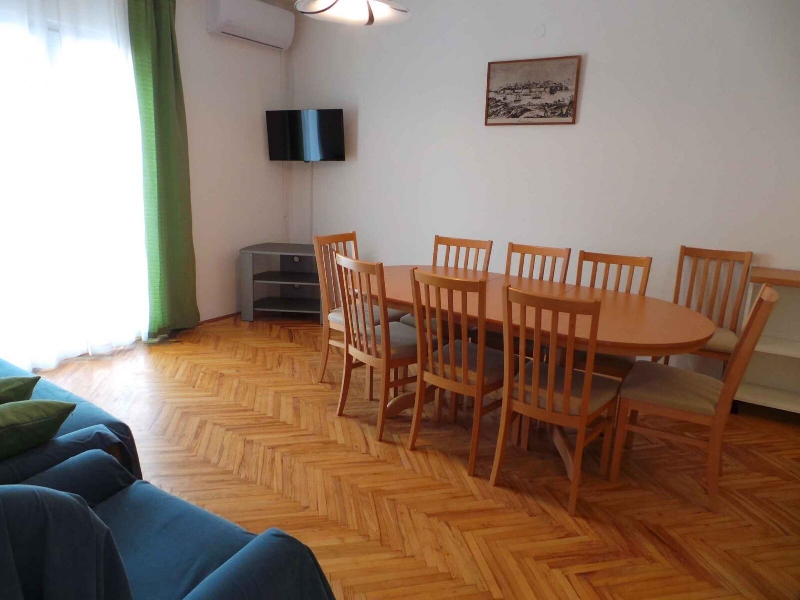 A dining area with a wooden table and chairs, a television mounted on the wall, and a painting above the table.