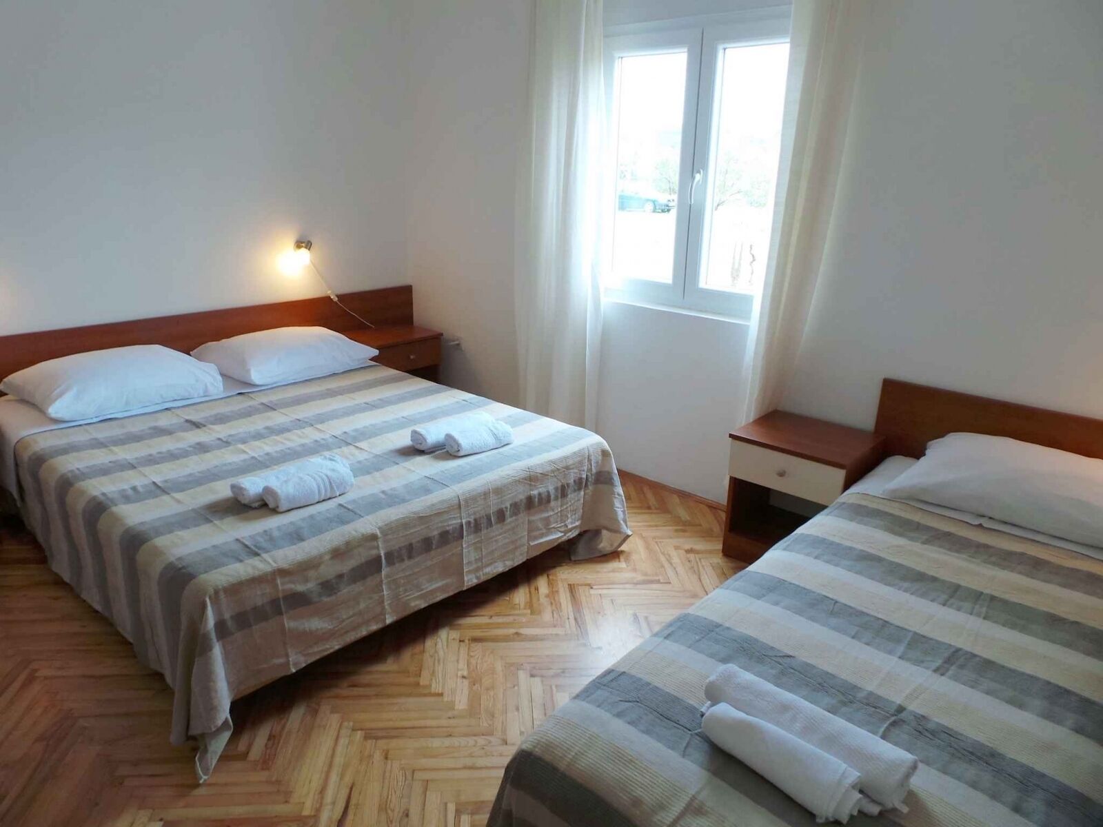 A hotel room with two beds, wooden flooring, a window with curtains, and a nightstand with a lamp.