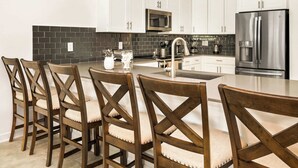 A modern kitchen with white cabinets, a stainless steel refrigerator, a microwave, and a sink. There are four wooden chairs with cushioned seats around a central island. - Luxe 8BR Villa by Disney | Arcade and Private Pool (Kissimmee)