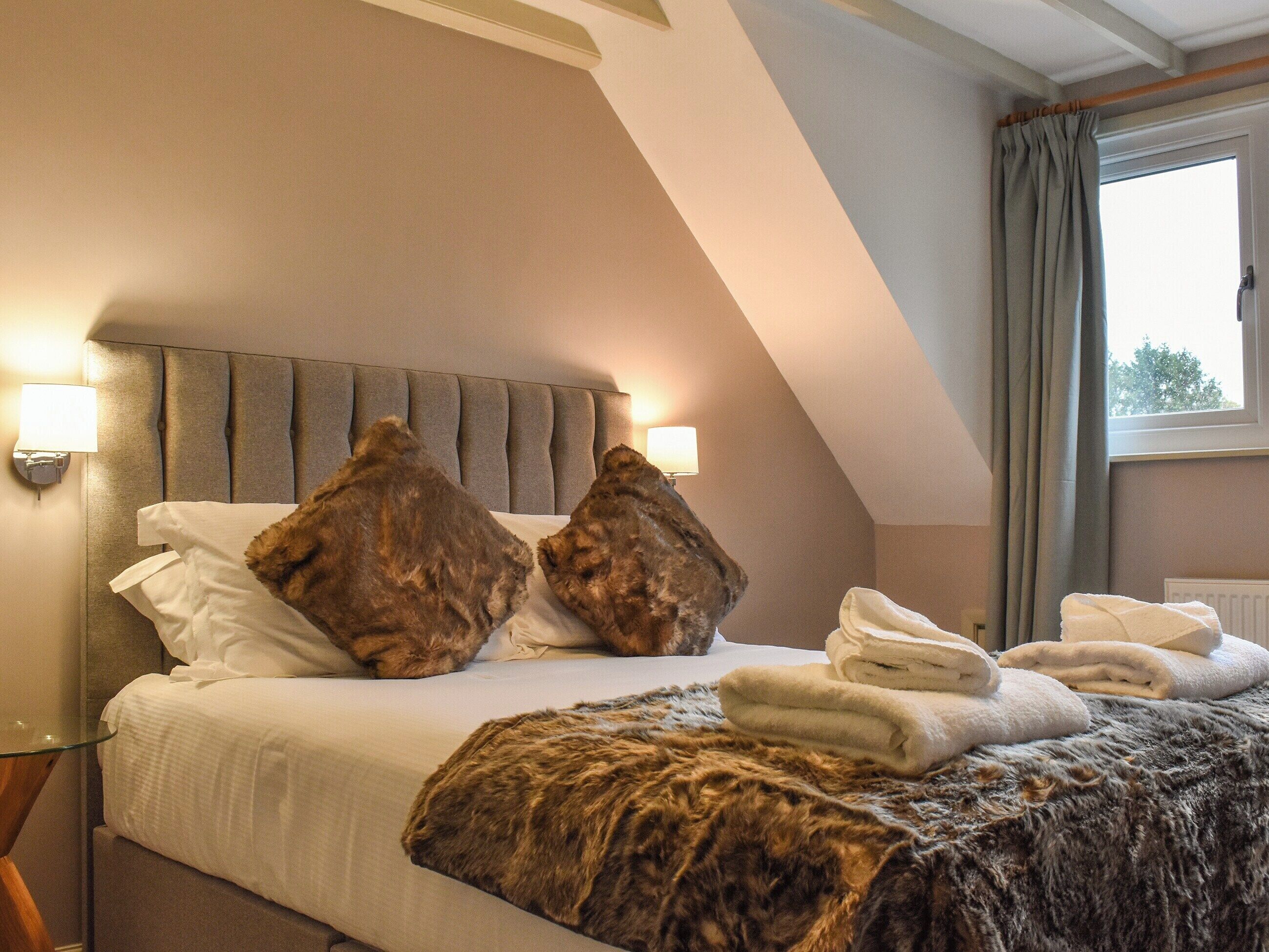 A hotel room with a bed, two pillows, a blanket, and a window with curtains.