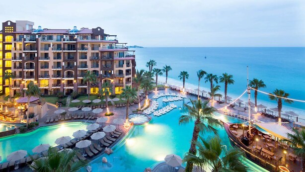 A hotel with multiple balconies, a large pool area with sun loungers and umbrellas, and a view of the ocean.
