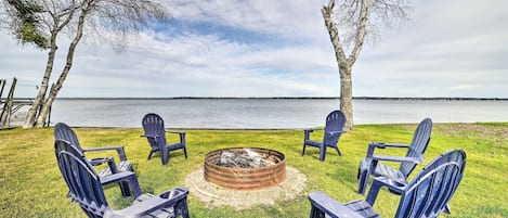 A fire pit surrounded by Adirondack chairs on a grassy area with a lake view.