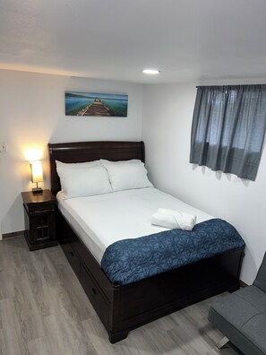 Una cama bien hecha con sábanas blancas y una manta azul estampada, una lámpara de noche, una mesita de noche de madera y un sofá gris, además de un cuadro de un muelle en la pared.