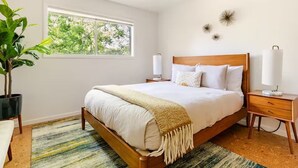 A bedroom with a wooden bed, bedside lamps, a potted plant, and a window with a view of trees. - Downtown Mid-Century Modern Condo with Retro Vibes (Boise)