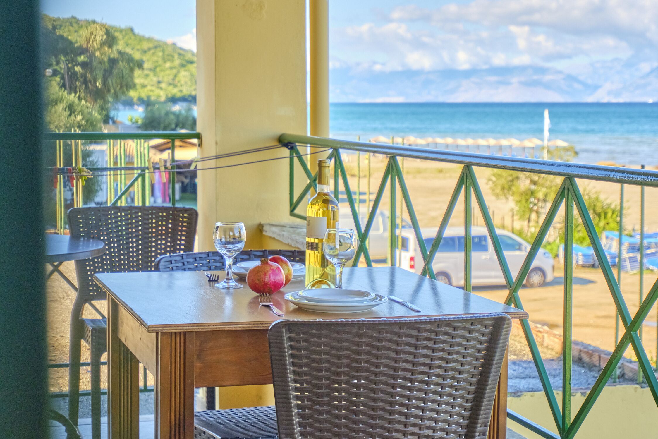 Ein Esstisch mit Stühlen, eine Weinflasche, eine Granatapfel und ein Glas Wasser auf einem Balkon mit Blick auf Strand und Meer.