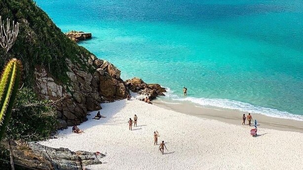 Une plage avec une eau turquoise et limpide, des affleurements rocheux et des gens qui se prélassent sur le sable.