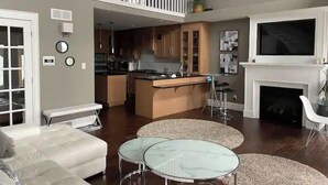 A spacious living area with a high ceiling, a kitchen with wooden cabinets, a dining area with a glass table, and a fireplace. - The Lakeside House With Hot Tub! (Amherstburg)