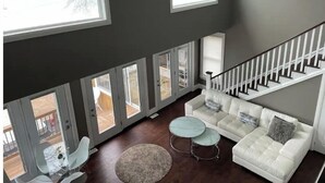 A spacious living area with a white sofa, two round coffee tables, and a view of a deck through large windows. - The Lakeside House With Hot Tub! (Amherstburg)