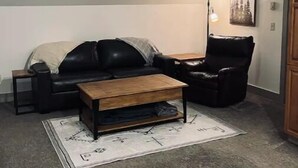 Ein Wohnzimmer mit einem schwarzen Ledersofa, einem Holztisch, einem schwarzen Leder-Sessel und einem kleinen Beistelltisch. An der Wand hängt ein gerahmtes Bild und es steht eine Stehlampe da.