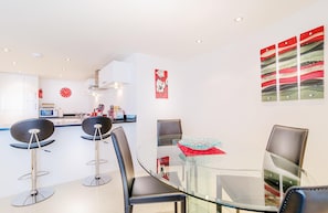 A modern kitchen with white cabinets, a microwave, and a red clock. There are two black bar stools and a glass table with chairs.