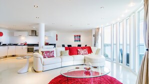 A modern living room with a white sofa, red accents, a glass coffee table, and a view of the city through large windows. - Spacious apartment, breathtaking sea views, private secure parking. (Newquay)