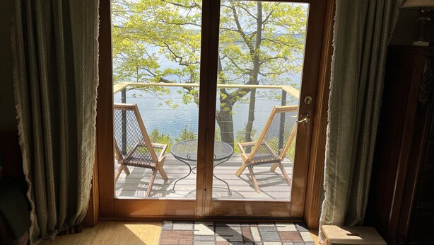 Een kamer met een schuifglazen deur die uitkomt op een terras met twee stoelen en een tafel, met uitzicht op een wateroppervlak en bomen.