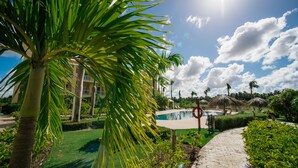 Zona de piscina en un resort, con cielo despejado, palmeras y un sendero pavimentado.