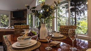 Een eetruimte met een houten tafel, geweven placemats, een vaas met groen en uitzicht op bomen door grote ramen.