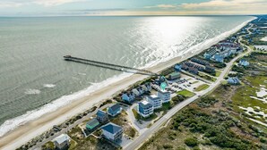 Vista aérea de una zona residencial costera con un muelle largo que se adentra en el mar, rodeada de casas de varios pisos y una carretera que discurre paralela a la playa.