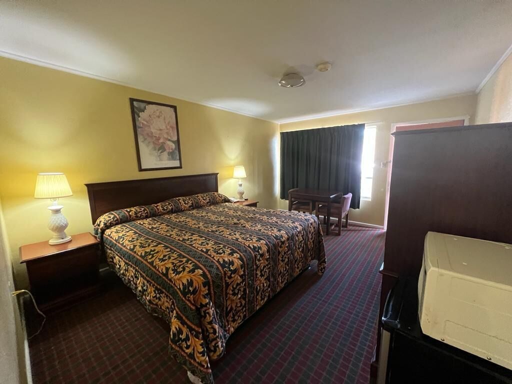A hotel room with a large bed, two bedside lamps, a desk with a chair, a large window with curtains, and a television.