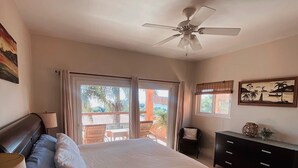 Een slaapkamer met een bed, een plafondventilator, een raam met gordijnen, een dressoir en een schilderij aan de muur.