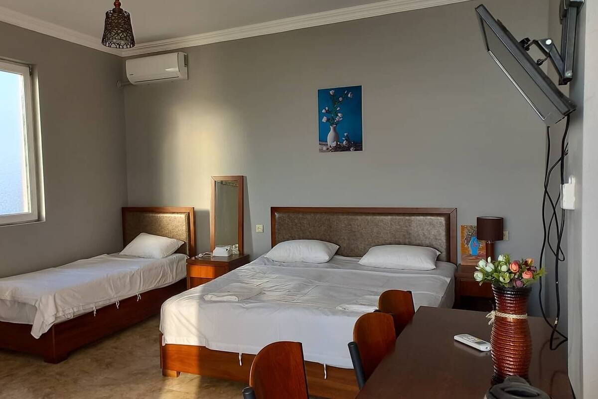 Habitación de hotel con dos camas individuales, una cama doble, un escritorio con sillas, una lámpara y un florero con flores.