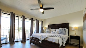 Un dormitorio con una cama grande, ventilador de techo y vistas al exterior a través de las ventanas.