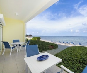 A balcony with a white table and blue chairs, overlooking a beach with lounge chairs and a clear sky.