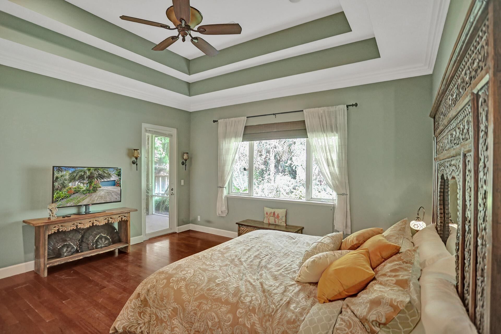 A bedroom with a bed, a ceiling fan, a window with curtains, a painting on the wall, and a wooden cabinet.