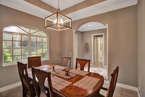 Une salle à manger avec une grande table, six chaises, un lustre et une vue sur un jardin grâce à de grandes fenêtres.