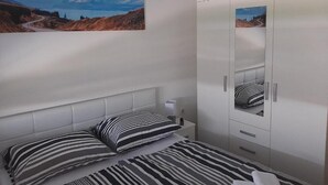 A bedroom with a bed, striped pillows, a nightstand, a wardrobe, and a painting of a mountainous landscape. - Apartment Kupre - One Bedroom Apartment with Sauna (Slavonski Brod)