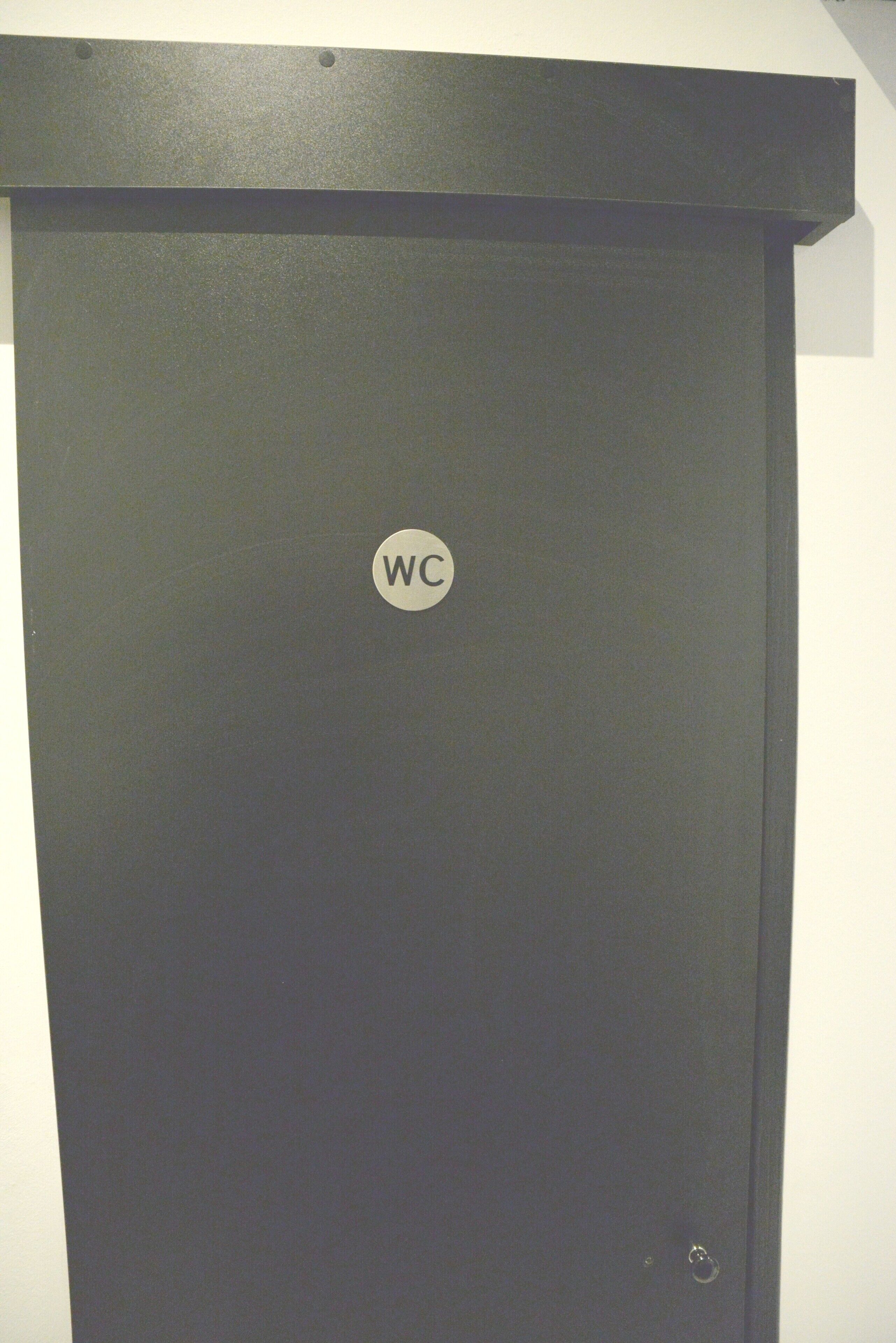 Ein dunkel gefärbtes, rechteckiges Schild mit der Aufschrift 'WC', das eine Toilette kennzeichnet.