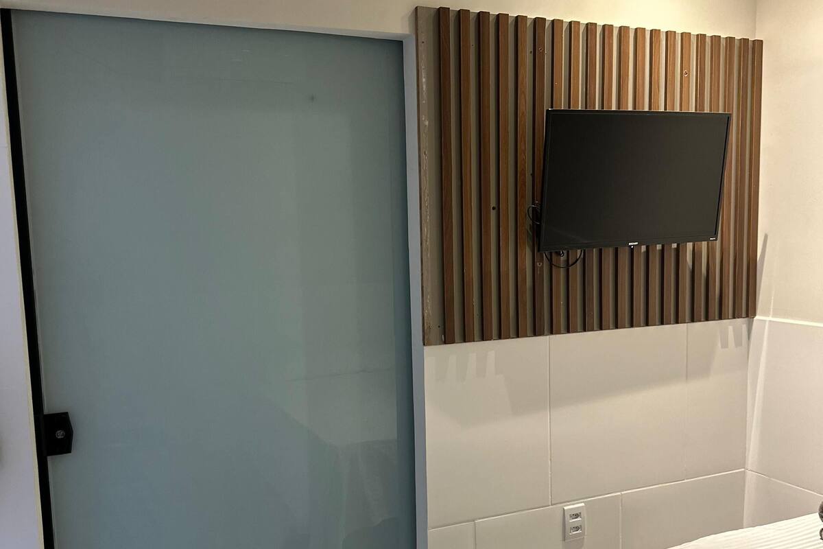 Quarto de hotel com porta deslizante, televisão de tela plana fixada em um painel de madeira e cama com colcha branca.