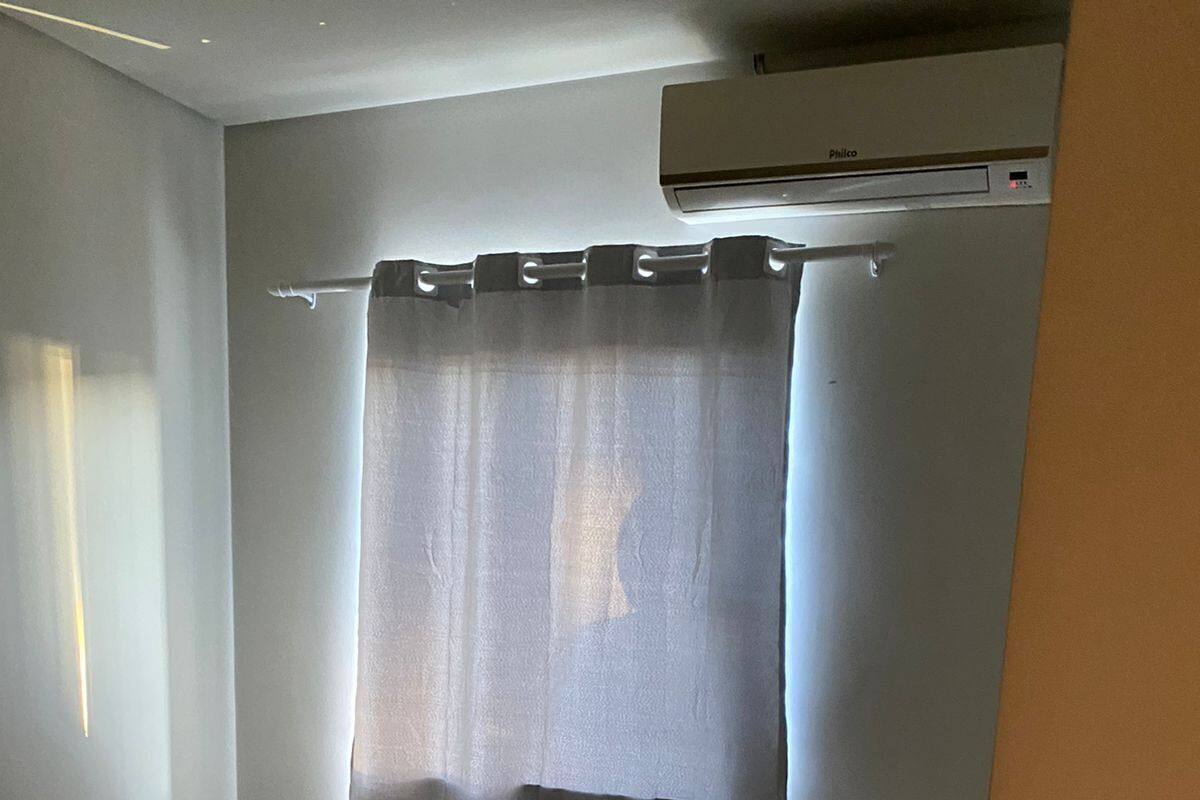 Quarto com cama, mesa de cabeceira, janela com cortinas e ar condicionado.