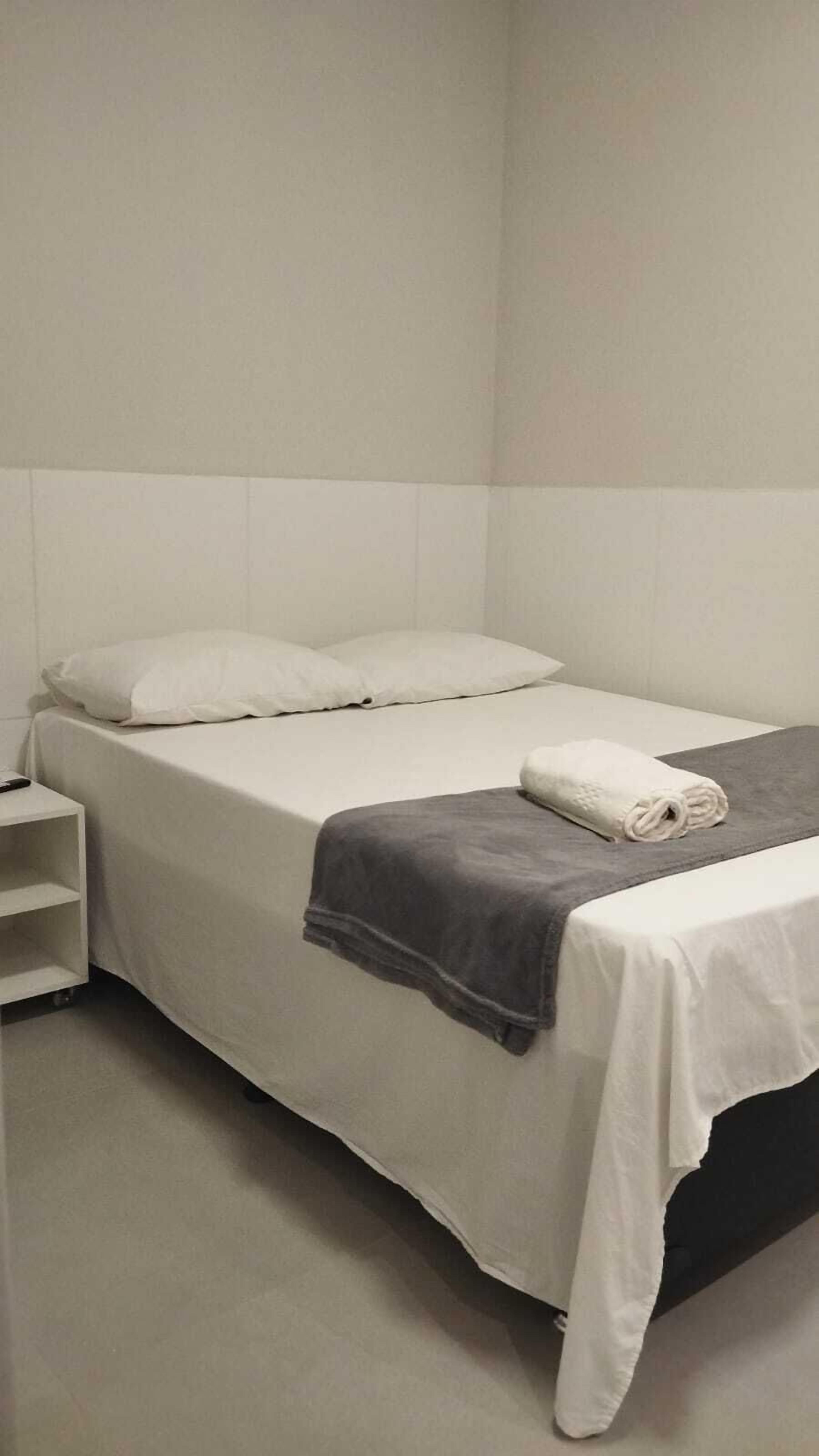 A single bed with white bedding, a grey blanket, and a rolled towel on a grey pillow.