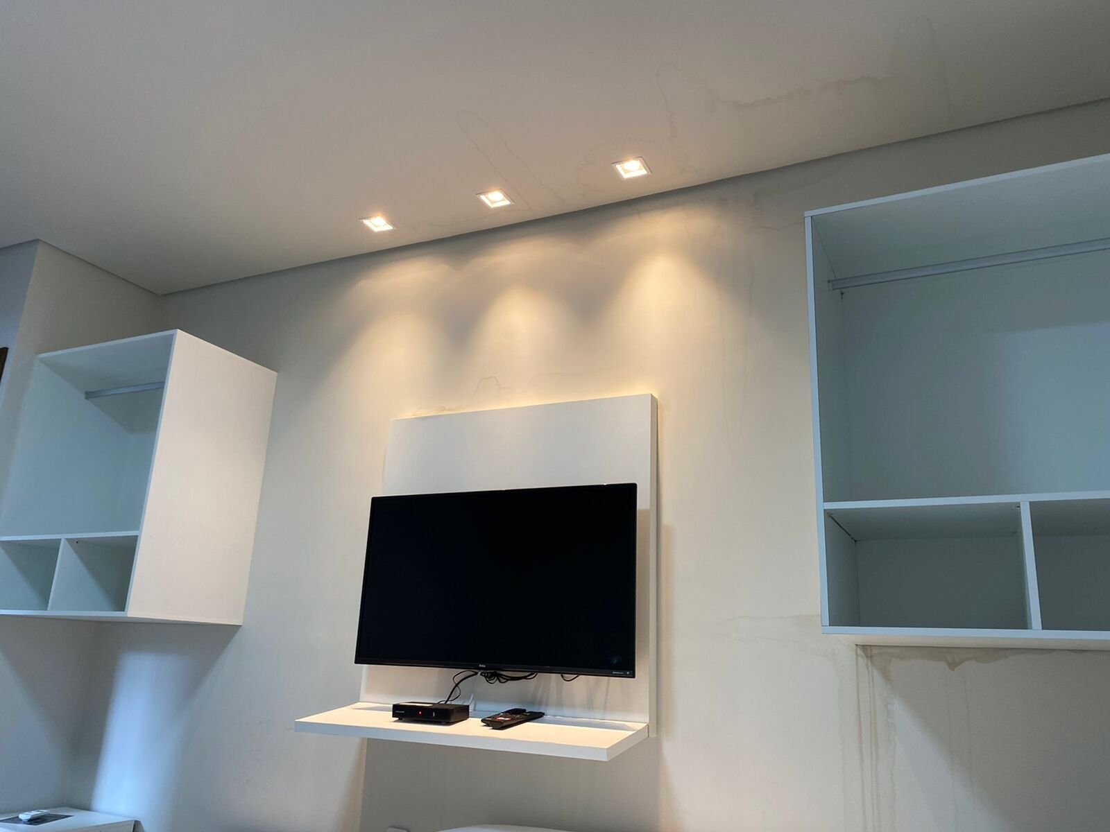 A modern bathroom with a mounted TV on a white shelf, recessed ceiling lighting, and built-in shelving units.