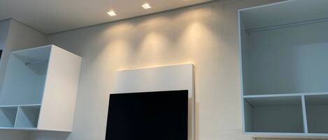 A modern bathroom with a mounted TV on a white shelf, recessed ceiling lighting, and built-in shelving units.