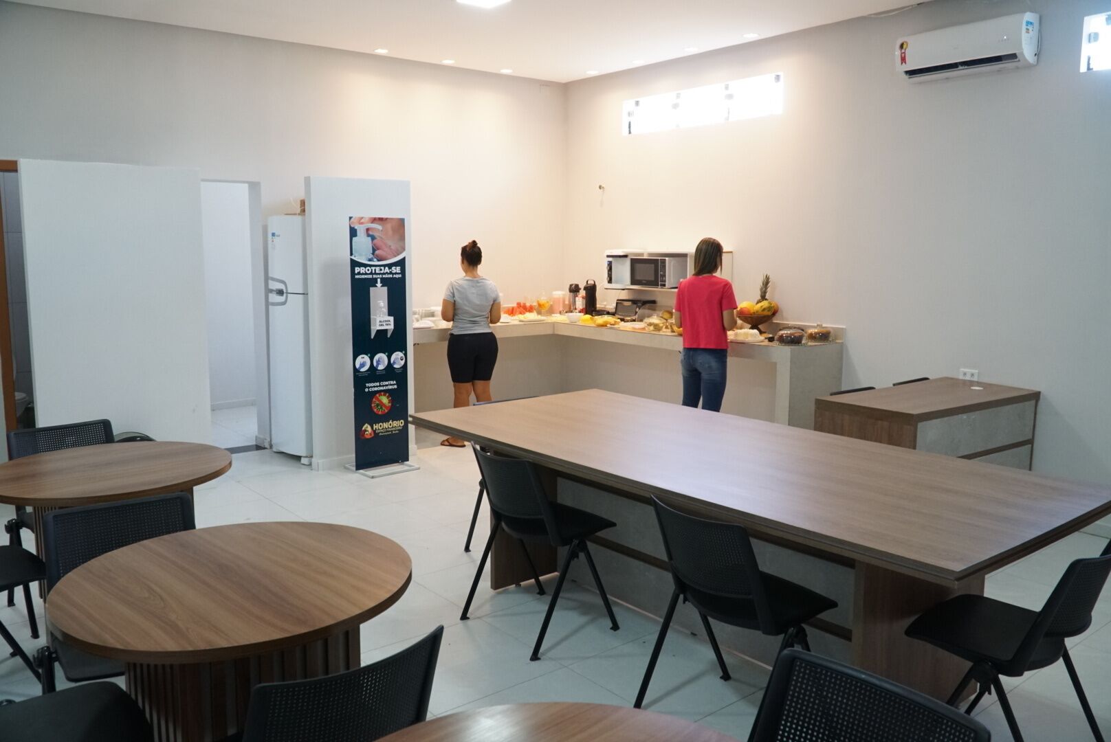A modern kitchen area with a microwave, refrigerator, and a person preparing food. There are tables and chairs arranged around the kitchen.