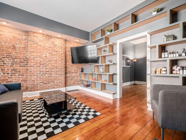 Una sala de estar moderna con pared de ladrillo, suelo de madera, alfombra a cuadros en blanco y negro, televisor de pantalla plana y estanterías empotradas con diversos objetos.
