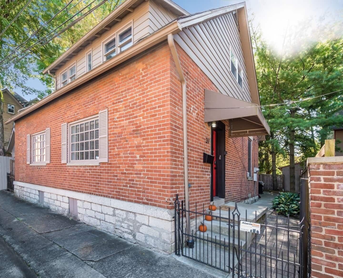 A two-story brick building with a red door and white shutters, featuring a small porch and a metal fence with pumpkins.