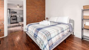 Un dormitorio con cama, suelo de madera, pared de ladrillo y cocina visible a través de un vano abierto.