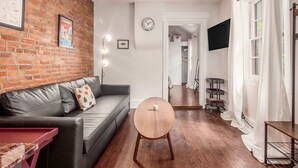 Una sala de estar moderna con una pared de ladrillo, un sofá gris, una mesa redonda de madera y una puerta blanca que da a otra habitación.