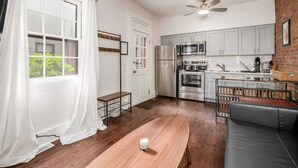 Una cocina moderna con electrodomésticos de acero inoxidable, una zona de comedor con una mesa de madera y un sofá gris, y una pared de ladrillo.