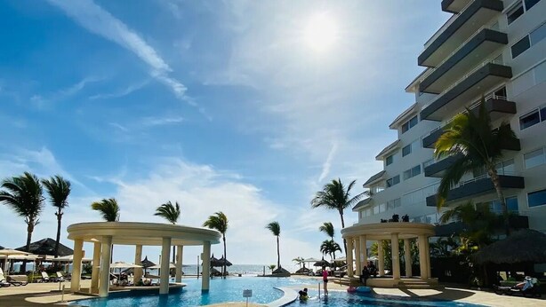 A large outdoor swimming pool with a clear blue sky, palm trees, and a multi-story building in the background. - Departamentos QM1 (Mazatlán)