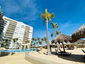 A resort with a swimming pool, palm trees, and a large hotel building. - Departamentos QM1 (Mazatlán)