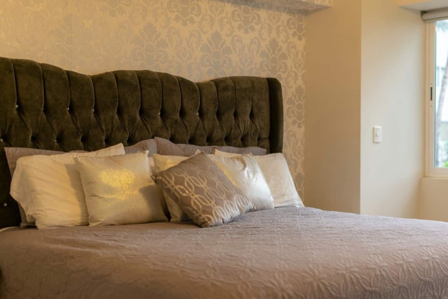 Una cama con cabecera capitonada, varios almohadones y un papel tapiz estampado.