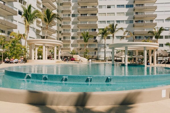 A large pool with multiple water features, surrounded by palm trees and a multi-story hotel building with balconies. - Departamentos QM1 (Mazatlán)