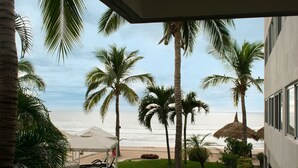 A balcony with a view of the beach, palm trees, and a thatched umbrella. - Departamentos QM1 (Mazatlán)