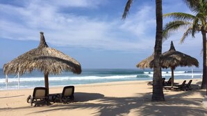A beach with two thatched umbrellas and lounge chairs, palm trees, and the ocean in the background. - Departamentos QM1 (Mazatlán)